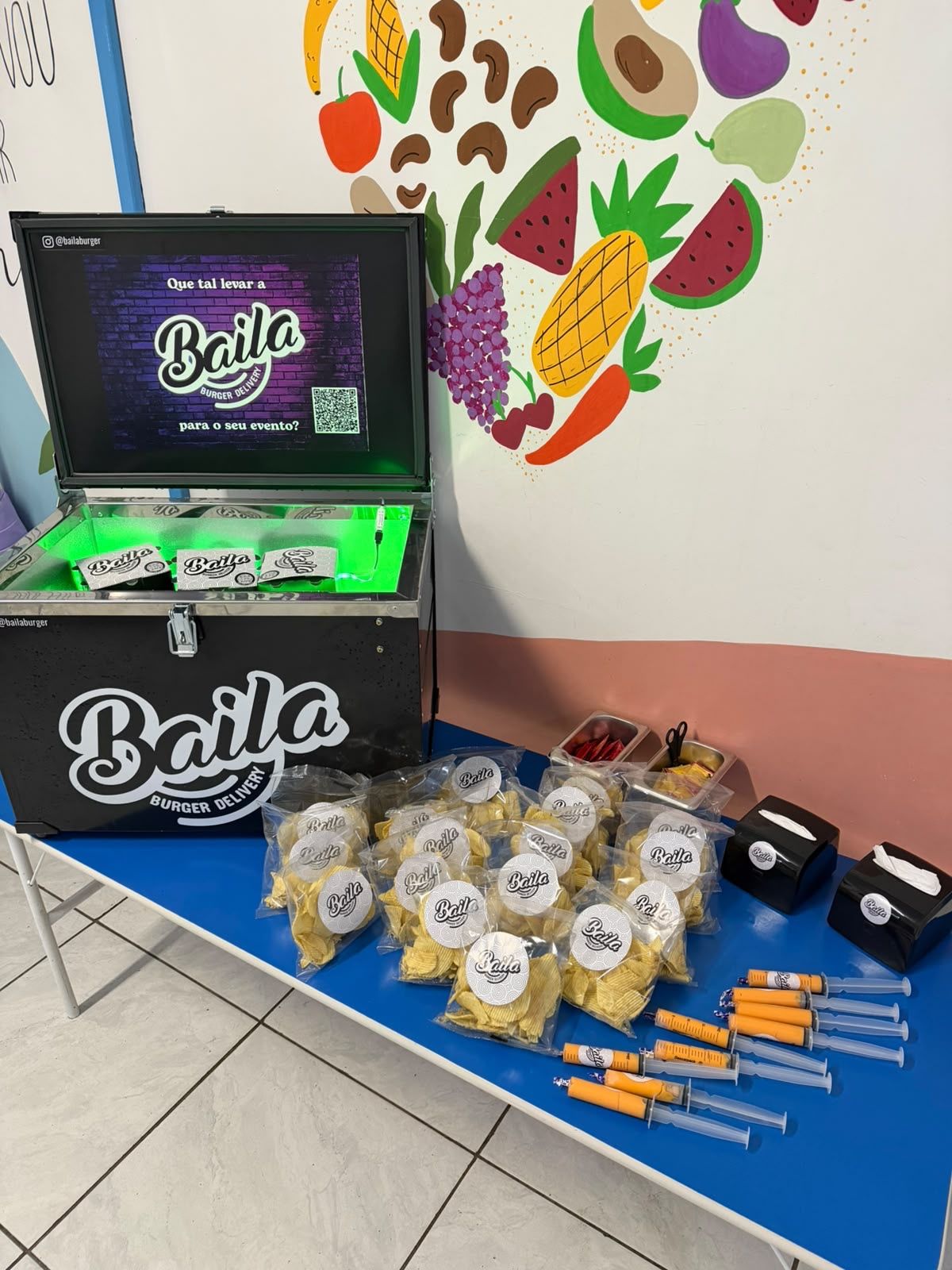 Estande completo Baila em evento, com tela exibindo QR code, saquinhos de chips personalizados e seringas de molho cheddar