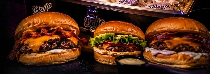 Três burgers artesanais Baila lado a lado em cima de uma mesa escura, com a box térmica ao fundo